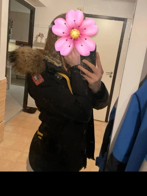 Svart parkajacka med päls från Parajumpers - Säljer en svart parkajacka från Parajumpers med stor fluffig päls på huvan och snygga gula detaljer. Jackan har lång ärm, patch på axeln och dragkedja framtill. Perfekt för kalla dagar och riktigt stilren look.