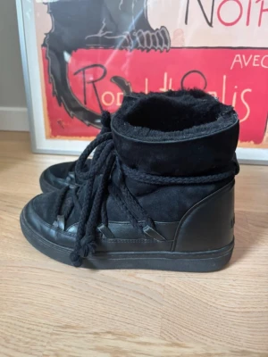 Svarta Inuikii boots - Säljer ett par svarta boots med snörning och mjukt pälsfoder på insidan. Skorna har en platt sula och är tillverkade i en kombination av mocka och skinn, vilket ger dem en snygg och modern look. Perfekta för kyliga dagar och passar till många olika stilar.