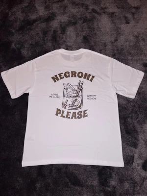 ”Negroni please” T-shirt - Vit t-shirt med stilrent tryck av ett Negroni-glas och texten “Negroni Please – Leave me alone with my Negroni”. En humoristisk och snygg tröja för drink- och cocktailälskaren, perfekt till vardags eller fest