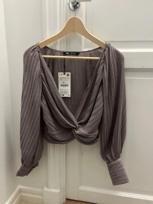 Plisserad omlottopp från Zara - Säljer en snygg plisserad omlottopp från Zara i en grå nyans. Toppen har långärmade ballongärmar med breda muddar och ett twistat omlottparti framtill. Helt ny!.