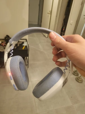 Logitech G435 Wireless Gaming Headset - Logitech G435 trådlöst gamingheadset i vitt och ljusblått. Lätt design, bekväma öronkuddar och tydliga knappar. USB-dongel medföljer för enkel anslutning. Headsetet ser ut att vara i gott skick utan synligt slitage eller defekter.