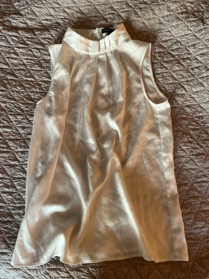 Vit ärmlös blus från STOCKH LM - Säljer en elegant vit blus från STOCKH LM i storlek S. Blusen är ärmlös med hög krage och har snygga veck framtill. Materialet är glansigt och känns lyxigt, troligen polyester eller satin. Dragkedja i nacken för enkel påklädning. Perfekt för dig som gillar stilrena plagg.