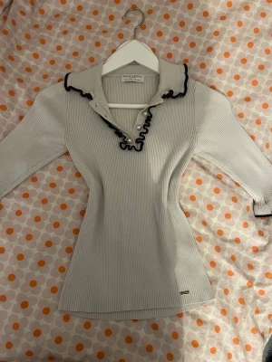 Vit ribbad topp från Holly & Whyte - Supersöt vit ribbad topp från Holly & Whyte med svarta volangdetaljer och pärlknappar vid kragen, kommer även med en extra knapp. Toppen har trekvartsärm och en figurnära passform. Perfekt för dig som gillar klassiska men ändå unika detaljer. Den har bara använts få gånger. 💕priset kan diskuteras 