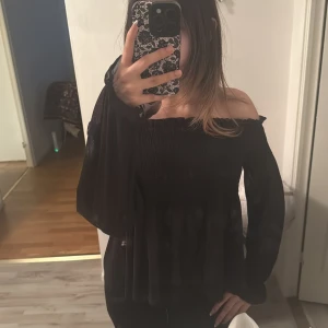 Svart offshoulder topp från Gina Tricot - Säljer en svart offshoulder topp från Gina Tricot i storlek S. Toppen har smockad överdel, långa ärmar med resår vid ärmslut och är tillverkad i ett lätt, transparent syntetmaterial. Perfekt för dig som gillar en trendig och luftig look.