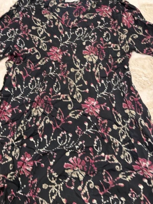 Blommig klänning från Masai XXL - Snygg svart klänning från Masai i storlek XXL med blommigt mönster i vinrött och beige. Klänningen har en mjuk och luftig känsla, perfekt för dig som gillar färg och mönster. Passformen är loose och materialet känns som viskos eller polyester.