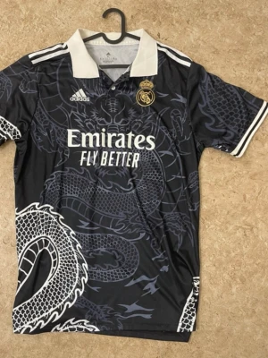 Svart Real Madrid tröja med drakmönster - Säljer en svart Real Madrid tröja från Adidas med ett fett drakmönster över hela plagget. Tröjan har vit krage och vita detaljer på ärmarna, samt klubbmärke och Adidas-logga på bröstet. Materialet är polyester och trycket är riktigt snyggt och unikt.