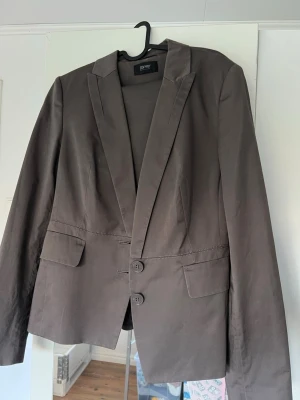 Brun kavaj från Esprit Collection - Snygg brun kavaj från Esprit Collection i klassisk modell med två knappar framtill och fickor med lock. Kavajen har långärmade ärmar och är tillverkad i ett mjukt bomullsmaterial. Perfekt för dig som vill ha en stilren och enkel look.