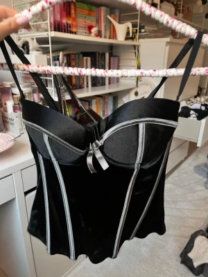 Svart sammets-korsett med silverdetaljer - Supersnygg svart korsettopp i sammet med silvriga sömmar och en liten rosett framtill. Justerbara axelband och hakknäppning bak för perfekt passform. perfekt fest topp!