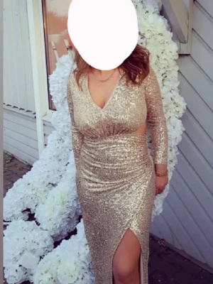 Glittrig långklänning med slits - Säljer en glittrig långklänning i guld med v-ringad hals och långärmad design. Klänningen har en snygg slits framtill och är figurnära med paljettdetaljer som ger extra glamour. Perfekt för speciella tillfällen och fester.