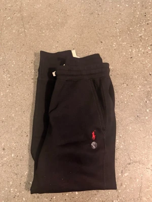 Svarta mjukisbyxor från Polo Ralph Lauren - Svarta mjukisbyxor från Polo Ralph Lauren med röd broderad logga på benet och vit snörning i midjan. Byxorna har ribbade muddar vid bensluten och är gjorda i mjukt bomullsmaterial. Perfekta för chill eller häng med kompisar. Ny pris 1995
