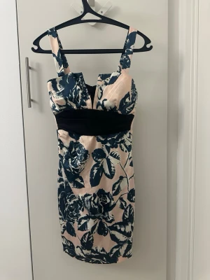 Blommig kort klänning med svart midja - Snygg kort klänning med breda axelband och blommigt mönster i mörkblått och ljusrosa. Klänningen har en markerad svart midja och en cool v-ringad detalj framtill. Materialet känns glansigt och har en lyxig känsla, perfekt för dig som vill sticka ut.