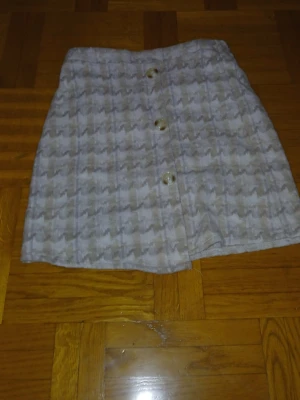 Beige och vit rutig kortkjol - Säljer en beige och vit kortkjol med rutigt mönster och knappar framtill. Kjolen har en mjuk och lite fluffig känsla, med resår i midjan för extra komfort. Perfekt för dig som gillar klassisk stil med en twist.