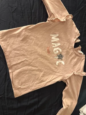 Beige långärmad topp från MNG - Säljer en beige långärmad topp från MNG med texten 'MAGIC Touch!' framtill och en glittrig stjärna i paljetter. Toppen har volangdetaljer vid axlarna och är tillverkad i mjuk bomull. Perfekt för dig som gillar gulliga detaljer och bekväma plagg.