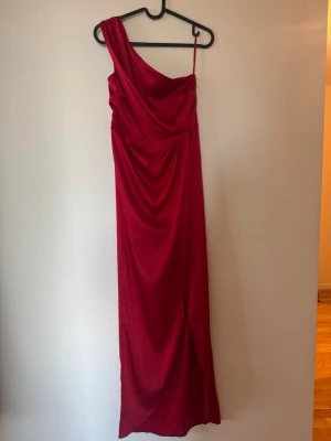 Röd one shoulder långklänning Nelly - Säljer en elegant röd långklänning från Nelly i storlek 36. Klänningen har one shoulder-design med smal axelrem och draperad omlott framtill. Använd 1 gång och i jättefint skick.