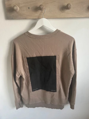Beige sweatshirt från Zara - Säljer en beige sweatshirt från Zara med rund hals och ribbade muddar. Tröjan har ett stort svart fyrkantigt tryck på ryggen och diskret broderad text framtill. Perfekt för dig som gillar stilrena detaljer och avslappnad passform.