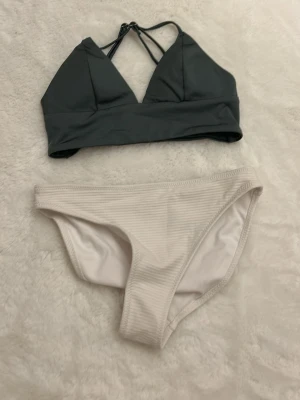 Mörkgrå bikiniöverdel från H&M - Snygg bikiniöverdel i mörkgrå färg med smala axelband som korsas i ryggen. Materialet är mjukt och stretchigt, perfekt för strandhäng. Matchas med ribbade vita bikinitrosor i samma storlek från H&M. Båda delar är tillverkade i polyester och elastan.🥰