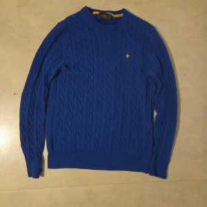 Blå kabelstickad tröja från Morris - Säljer en blå kabelstickad tröja från Morris i storlek L, tillverkad av 100% pima bomull. Tröjan har rund hals, ribbade muddar och ett litet broderat emblem på bröstet. Perfekt för dig som gillar klassisk stil med en modern touch.