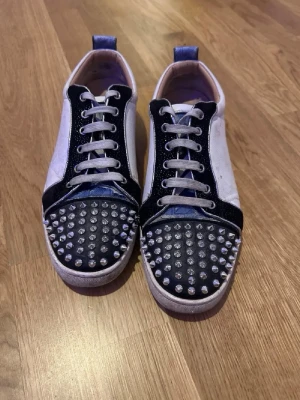 Unika loubs - Säljer ett par unika sneakers från Christian Louboutin med svart och vit färg, dekorerade med silvriga nitar på tån. Skorna har snörning, rund tå och röd sula med logotyp. Materialet är skinn och textil, och de har en cool kontrast mellan färgerna.