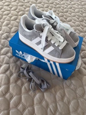 Adidas Campus 00s grå sneakers - Adidas Campus 00s sneakers i grå mocka med vita detaljer och klassiska tre ränder på sidan. Skorna har vit snörning, rund tå och platt sula. Medföljer extra grå skosnören. Perfekt för dig som gillar retrostil och streetwear.