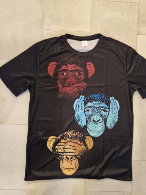 Svart t-shirt med aptryck - Cool svart t-shirt i polyester med tryck av tre apor i rött, blått och gult på både fram- och baksidan. Polyester. T-shirten har rund hals och korta ärmar, perfekt för dig som gillar unika och färgstarka motiv.