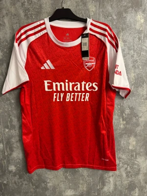 Arsenal Saka - Ny 2XL - Bilderna visar en röd och vit Arsenal fotbollströja med Saka 7 på ryggen, tillverkad av Adidas. Ingen sko finns med på bilderna.