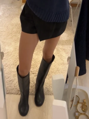 Svarta höga boots i skinn - Säljer ett par svarta boots med högt skaft och rund tå. Skorna är tillverkade i slätt skinn och har en platt sula som gör dem bekväma att gå i. Perfekta för dig som gillar stilrena och klassiska skor med lite edge.