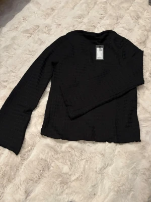 Svart långärmad topp med struktur - Säljer en svart långärmad topp från Lager 157 i storlek S. Toppen har en snygg strukturerad yta och är tillverkad i ett lätt syntetmaterial. Passformen är normal och toppen har en enkel, stilren design som funkar till många olika looks.