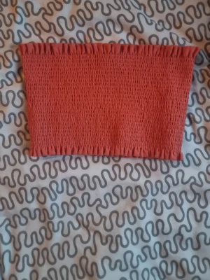 Rostfärgad bandeau tubtopp - Säljer en rostfärgad bandeau tubtopp med smockad struktur och elastiska kanter upptill och nedtill. Toppen är axelbandslös och har en tight passform som sitter snyggt mot kroppen. Perfekt för varma dagar och sommarhäng.