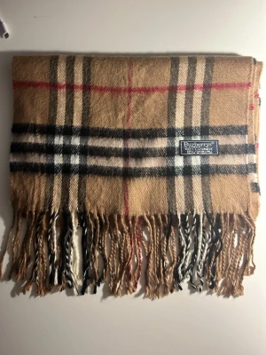 Rutig beige väst från Burberrys - Säljer en klassisk rutig väst från Burberrys i 100% kashmir. Västen har beige bas med svarta, vita och röda rutor, samt fransar längs nederkanten. Perfekt för dig som gillar tidlös design och mjuka material.