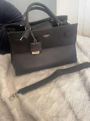 Svart handväska från Guess - Säljer en stilren svart handväska från Guess med guldfärgade detaljer och logotyp framtill. Väskan är tillverkad i strukturerat skinn och har både handtag och avtagbar axelrem. Insidan är fodrad med mönstrat tyg och har flera fack med dragkedja för smart förvaring.