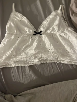 Vit broderad linnetopp med rosett - Säljer en söt vit linnetopp med broderade blommor och spetskant vid urringningen. Toppen har smala axelband och en liten svart rosett framtill. 