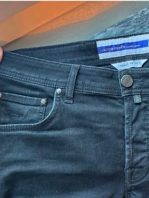 Svarta jeans från Jacob Cohen Nick - Snygga svarta jeans från Jacob Cohen, modell Nick. Jeansen har klassisk femficksdesign, silverfärgade knappar med logga och broderad J på fickan. Slimmad passform och tillverkade i mjukt bomullsmaterial med stretch för extra komfort. Made in Italy.