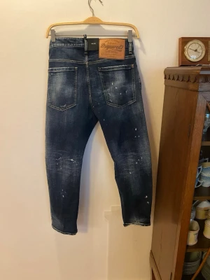 Dsquared2 mörkblå croppade jeans - Säljer ett par mörkblå jeans från Dsquared2 med croppad passform och coola slitningar samt vita färgstänk. Klassisk femficksmodell med läderpatch bak i midjan och plomberingen sitter kvar. Perfekt för dig som gillar streetstyle och vill sticka ut.