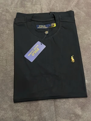 Svart t-shirt från Polo Ralph Lauren - Klassisk svart t-shirt från Polo Ralph Lauren med gul broderad logga på bröstet. T-shirten har rund halsringning och korta ärmar. Tillverkad i mjuk bomull för en skön känsla och clean look. Perfekt till jeans eller shorts.