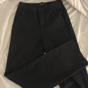 Svarta kostymbyxor från Vero Moda - Snygga svarta kostymbyxor från Vero Moda med klassisk rak passform och medelhög midja. Byxorna har sidfickor och stängs med dragkedja och knapp. 