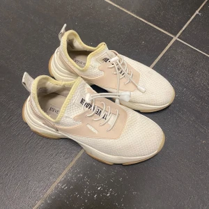 Steve Madden vita sneakers med beige detaljer - Snygga sneakers från Steve Madden i vit mesh med beige och ljusgula detaljer. Skorna har rund tå, platt sula och snörning med snabblås. Yttermaterialet är syntet och textil, med en chunky sula i gummi. Perfekta för dig som gillar stilrena och moderna sneakers.