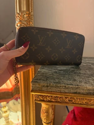 Monogram plånbok - Säljer en klassisk brun plånbok från med ikoniskt monogrammönster och guldfärgad kedjedetalj. Plånboken har dragkedja runtom och flera fack inuti för kort och kontanter. Perfekt accessoar för dig som gillar lyxig och tidlös design.