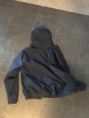 Mörkblå vindjacka från Lyle & Scott - Snygg mörkblå vindjacka från Lyle & Scott med huva och dragkedja framtill. Jackan har ett stilrent utseende med gul logga på bröstet och ribbade muddar vid ärmslut och nederkant. Perfekt för dig som vill ha en enkel och cool jacka.