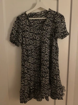 Svartvit kortklänning Vero Moda - Kort klänning från Vero Moda AWARE i svartvitt abstrakt mönster. Modellen har korta puffärmar och rund halsringning. Tillverkad i ett lätt och luftigt material, perfekt för varmare dagar. Snygg och enkel att styla med sneakers eller boots.