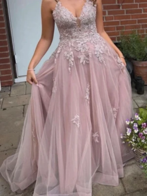 Ljusrosa balklänning med spetsdetaljer - Romantisk ljusrosa/smutsrosa balklänning med vacker spets och blommiga applikationer. Klänningen har en djup urringning, tunna axelband som korsas i ryggen och en vid, svepande kjol i tyll. Öppen rygg med snörning för justerbar passform. Perfekt för bal eller speciella tillfällen.
