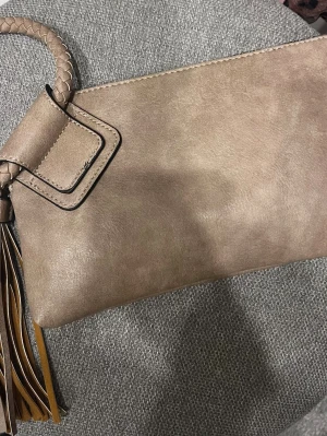 Beige kuvertväska med flätad handledsrem - Stilren beige kuvertväska i läderimitation med flätad handledsrem och dekorativ tofs. Väskan har dragkedja upptill och en enkel, elegant design som passar till många olika looks. Perfekt för dig som gillar minimalistisk stil.
