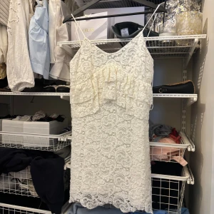 Vit spetsklänning med volang - Söt vit kortklänning i spets med tunna axelband och volangdetaljer framtill. Klänningen har en figurnära passform och vacker struktur i spetsmaterialet. Perfekt för sommarens alla tillfällen.