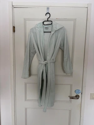 Ljusblå/turkos fluffig morgonrock med huva - Mysig ljusblå morgonrock i supermjukt fleece-material. Har huva med två fluffiga bollar baktill och knytskärp i midjan. Långa ärmar och enkel, avslappnad passform. I storlek 146/152 från Kappahl. Perfekt för chilliga morgnar eller kvällar hemma. Skicka för fler bilder eller annat.