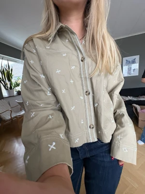 Beige overshirt jacka med broderi - Trendig beige overshirt jacka i bomull med vita broderade kryssdetaljer över hela plagget. Jackan har knappar framtill, croppad passform och långa ärmar. Perfekt för lager på lager under vår och höst.