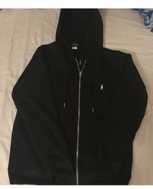Svart hoodie från Polo Ralph Lauren - Säljer en svart hoodie från Polo Ralph Lauren med dragkedja och vit broderad logga på bröstet. Hoodien har huva med dragsko och två fickor framtill. Klassisk och stilren design i mjukt material, perfekt för en avslappnad look. Jag har aldrig andvänt den innan 