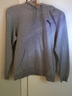 Grå hoodie från Puma med logga - Säljer en klassisk grå hoodie från Puma med svart logga på bröstet. Hoodien har huva med dragsko och en stor magficka framtill. Tillverkad i mjukt material som passar perfekt för chill dagar. Enkel och stilren design som funkar till det mesta.