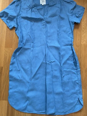 Blå kort klänning från Part Two - Ljusblå klänning från Part Two i mjukt linnematerial. Klänningen har korta ärmar, v-ringad hals och avslappnad passform. Perfekt för varma dagar och enkel att styla med accessoarer.