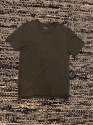 Mörkgrå t-shirt från BOSS - Snygg mörkgrå t-shirt från BOSS med diskret logga på bröstet. Klassisk rund halsringning och korta ärmar. Perfekt basplagg i garderoben och enkel att matcha med allt. Tillverkad i mjukt material för skön känsla hela dagen.