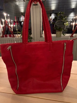 Röd handväska i skinn - Röd handväska i skinn från Ceannis med dekorerande dragkedjor på sidorna. Väskan har två handtag och stängs med dragkedja. Inuti finns ett fack med dragkedja och två öppna fack.  Mått 35x42x26 cm  Väskan är i mycket gott skick.