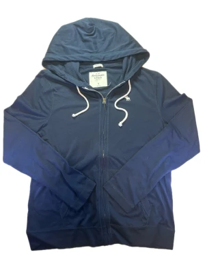 Marinblå hoodie från Abercrombie & Fitch - Snygg marinblå hoodie från Abercrombie & Fitch med dragkedja framtill och vita dragsnören i huvan. Tillverkad i mjukt material och har fickor på sidorna samt en liten broderad logga på bröstet. Perfekt för en avslappnad och stilren look.